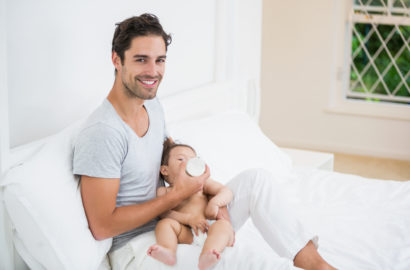 Father smiling on a sofa whilst bottle feeding a relaxed looking baby
