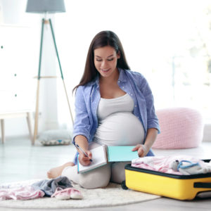 Pregnant mother smiling whilst sitting on the floor writing down a list with a suitcase half packed with baby things beside her