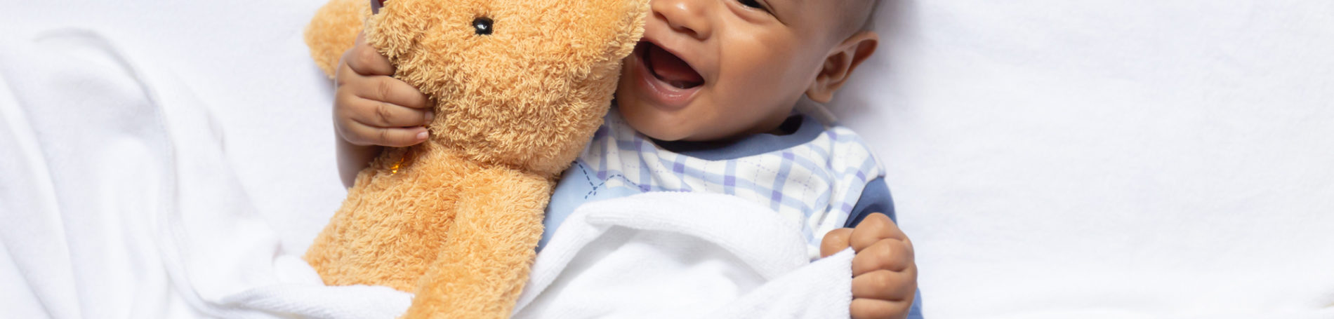 African baby laughing with a blanket over him and holding a teddy bear