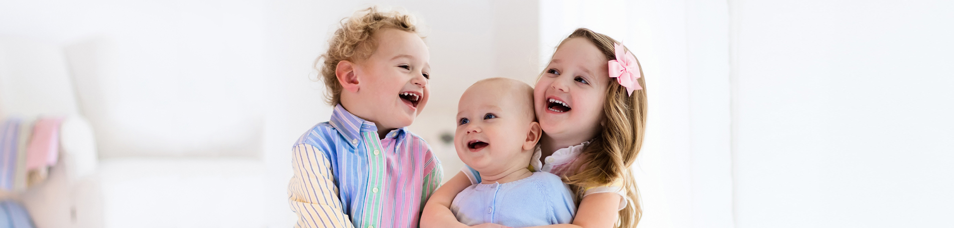 Two siblings, a girl and boy laughing whilst holding thier baby brother who is looking up a them and smiling