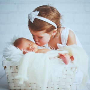 Toddler girl with a white hairband and a white dress kissing a very newborn looking baby who is sleeping on a fluffy white blanket in a crib
