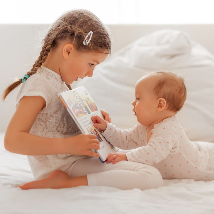 Lttle girl sitting on a bed and showing her baby sister the pictures in a book