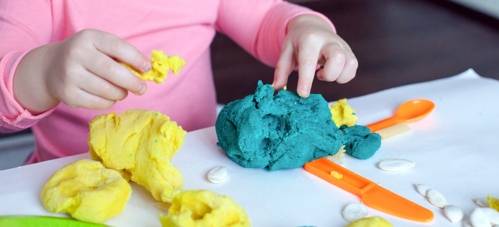 Childs hands playing with homemade green and yellow coloured playdough and a plastic knife and spoon on the table