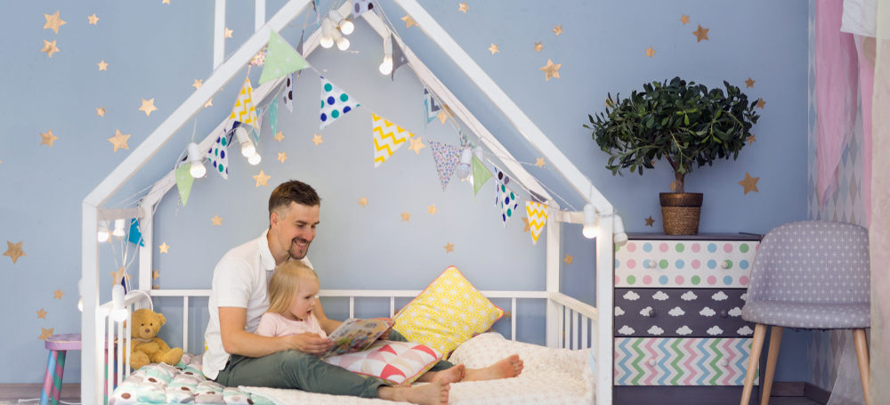 Father and 3 year old daughter in a framed toddler bed with the mattress at floor level. Stars on the wall and colourful flags attached to the frame of the bed which goes up and over the childs head in the shape of a house.