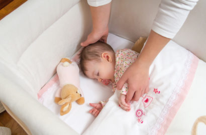 Baby in a crib with a dummy in her mouth and the mothers hands soothing her whilst she settles to sleep