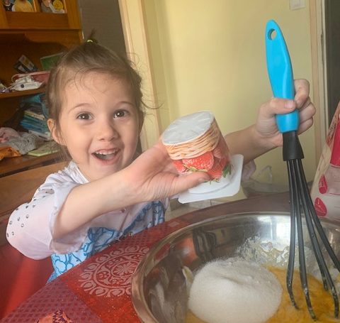 4 year old little girl looking excited whilst putting ingredients into a bowl with the use of a yogurt pot and a whisk in a bowl.